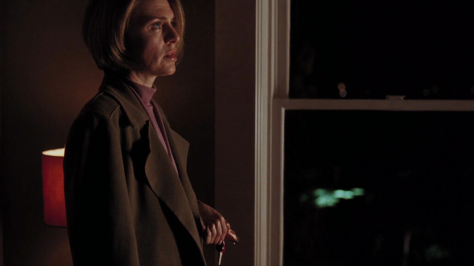 Una mujer con el abrigo puesto y las llaves en la mano, quieta junto a la ventana, de noche.