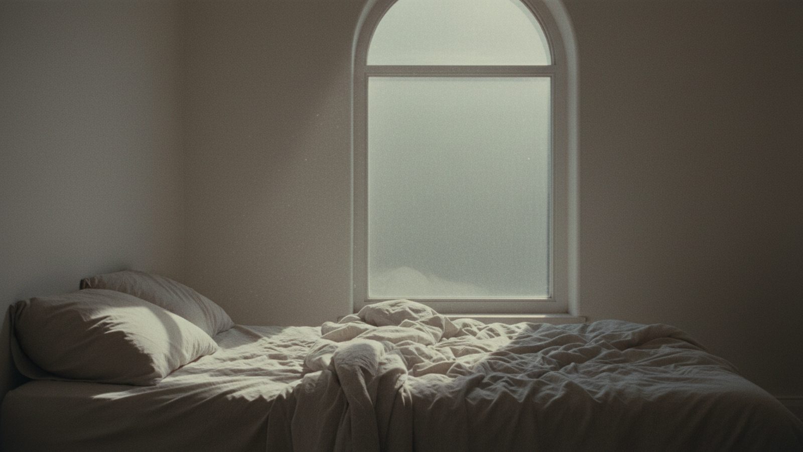 Una cama deshecha junto a una ventana, con la luz gris de una mañana avanzada.