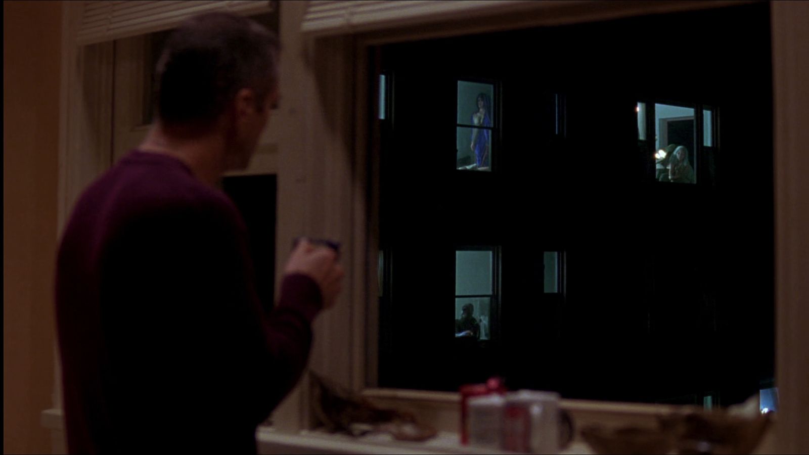 Un hombre mira por la ventana de su cocina, de noche, hacia las ventanas iluminadas de enfrente.