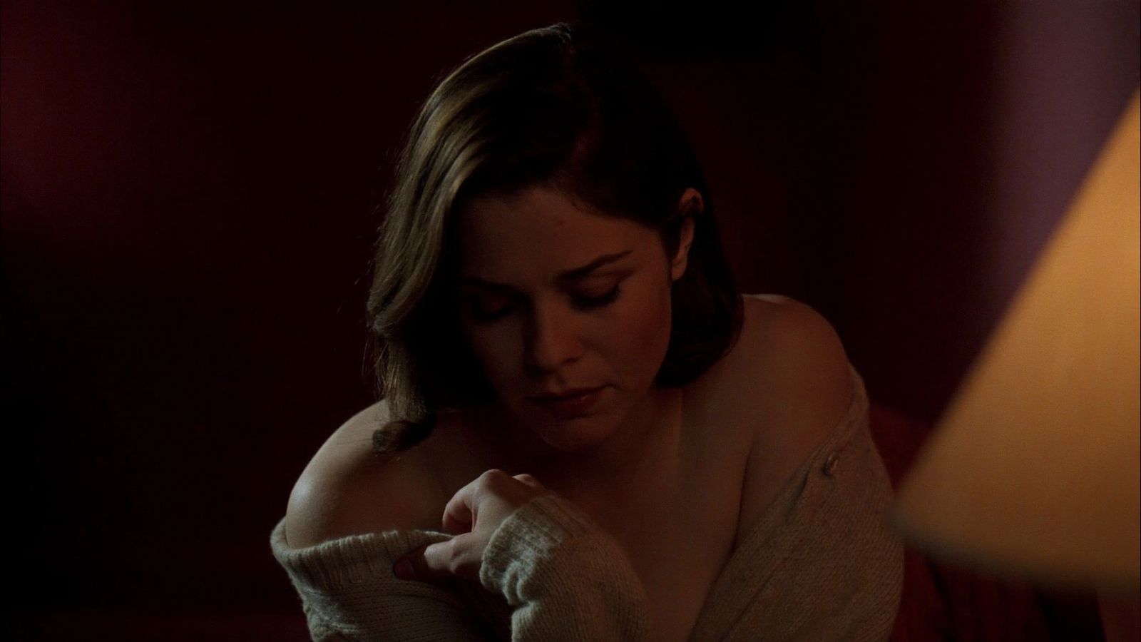 Una mujer sentada al borde de la cama, arropada en una chaqueta, la mirada baja.